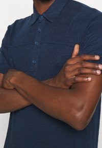 Námořnická polo košile vyrobená z texturované látky, s límečkem, knoflíkovou légou a krátkými rukávy. Viditelný detail na panelu v horní části hrudi.