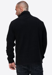 Czarna polarowa kurtka z wysokim kołnierzem, długimi rękawami i miękką teksturą. Posiada dyskretną metkę z logo na dolnym brzegu.