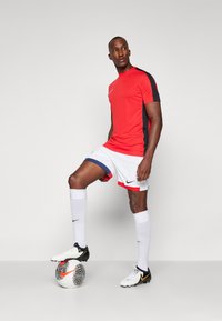 Rode en zwarte sporttrui met een ronde hals, gecombineerd met witte shorts met marineblauwe accenten. Model staat op een gemarmerde voetbalsbal.