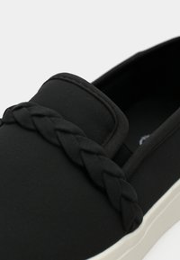 Black slip-on shoe with a textured canvas upper, featuring a braided accent on the vamp and a contrasting white rubber sole.