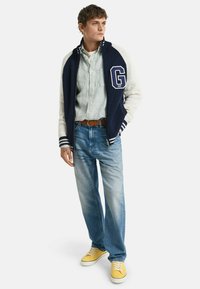Giacca varsity blu navy e crema con logo 'G', polsini a righe; camicia blu, jeans in denim chiaro e sneakers gialle completano l'outfit.