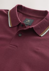 Bordeauxrotes Poloshirt aus strukturiertem Stoff, mit Kragen, zwei Knöpfen und einem gestreiften Kragen in Grau, Gelb und Weiß.