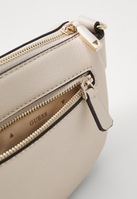 Beige kožená křížová taška se zlatými zipy. Disponuje texturovaným povrchem a několika oddíly, uvnitř je viditelné logo "GUESS".