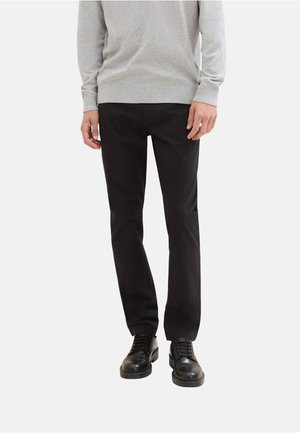 Hombre con pantalones negros, zapatos de cordones negros y un suéter de manga larga gris claro, de pie frente a un fondo blanco liso.