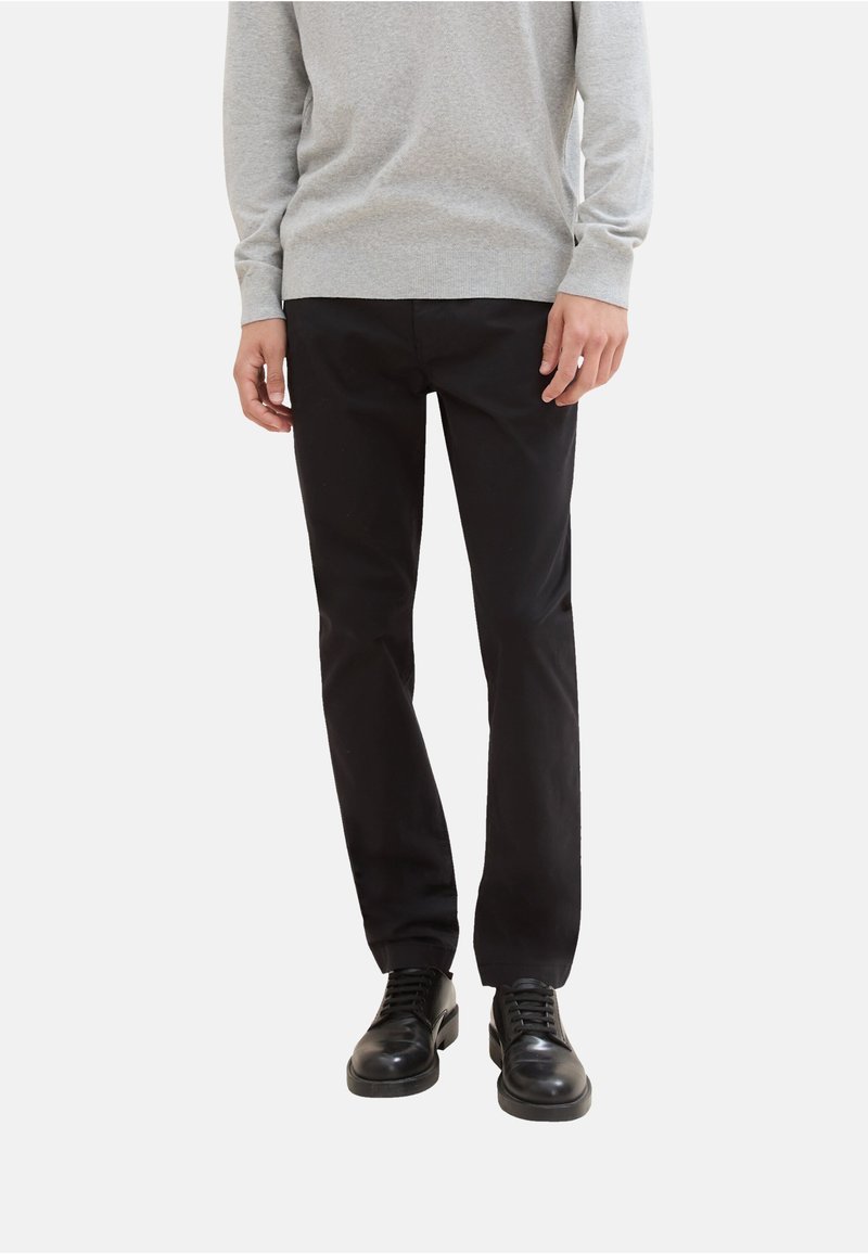 Man in zwarte broek, zwarte veterschoenen en een lichtgrijze lange mouwtrui, staande tegen een effen witte achtergrond.