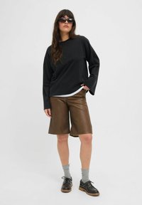 Melna garām piedurknēm krekls, brūni ādas šorti, pelēkas zeķes un melni apavi ar biezām zolitēm. Modelis stāv pret neitrālu fonu.