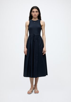 Γυναίκα με μακριά σκούρα μαλλιά που φοράει αμάνικο φόρεμα midi σε ναυτικό μπλε και λευκά σανδάλια, στέκεται μπροστά σε μια απλή ανοιχτή επιφάνεια.