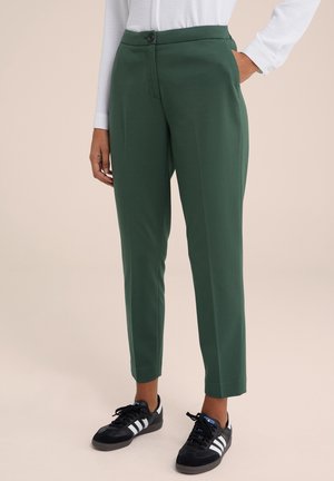 Pantalons ajustés verts avec une texture lisse, poches avant et fermeture à un bouton, associés à des chaussures de sport noires avec des rayures blanches.