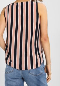 Donna che indossa un top senza maniche con strisce verticali nere, rosa e bianche e jeans di denim blu, vista da dietro su uno sfondo semplice.