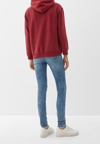 Sweat à capuche rouge avec une poche kangourou, associé à un jean skinny bleu clair. Des baskets blanches à semelle texturée complètent la tenue.