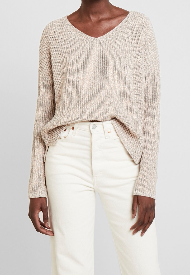 Pull beige en maille avec col en V, coupe ample et manches longues, associé à un jean blanc taille haute. Tissu texturé et doux.