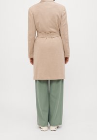 Abrigo beige con cinturón, de tela texturizada, de longitud a la rodilla, con una sola abertura en la parte posterior, combinado con pantalones verdes holgados y zapatos crema.