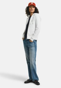 Witte zip-up hoodie met koordjes, gecombineerd met een marineblauw shirt en blauwe wijde jeans. Afgewerkt met zwarte schoenen en een rode pet met de letter "G."