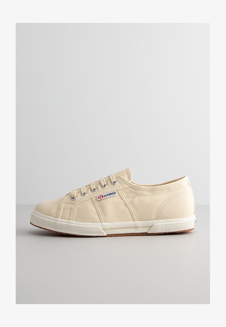 Beżowy płócienny sneaker niskoprofilowy z białą gumową podeszwą, okrągłym noskiem, sześcioma oczkami i widocznymi szwami. Znajduje się czerwono-niebieskie logo z boku.