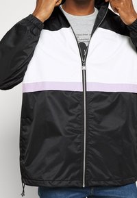 Schwarze, weiße und lavendelfarbene Windbreaker-Jacke mit Reißverschluss vorne, elastischen Bündchen und kontrastierenden Horizontalstreifen über der Brust.