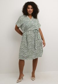 Vestido envolvente de manga corta en blanco con patrones abstractos en verde, cinturón a la cintura y tejido fluido. El dobladillo está por encima de la rodilla.