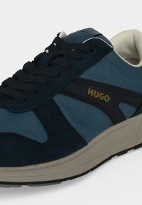 Baskets bleues et noires avec semelle beige, lacets noirs, logo doré "HUGO" sur le côté et col rembourré blanc.