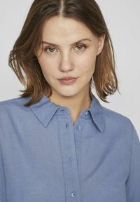 Svetlo modra srajca na gumb z mehko teksturo, s koničastim ovratnikom in sprednjim zapiranjem, ki vsebuje usklajene gumbe. Preprost in klasičen dizajn.