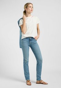 Camiseta blanca de manga corta con texto rosa, combinada con unos vaqueros de denim de color azul claro. El modelo lleva una chaqueta de denim colgada casualmente sobre un hombro.