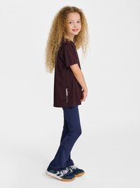 Ung pige med langt krøllet hår iført en mørk oversize t-shirt, marineblå leggings og marineblå sneakers med hvide snørebånd, står sideværts og smiler.