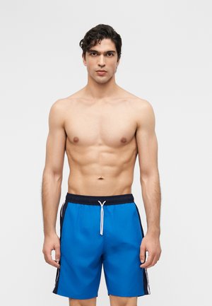 Jeune homme aux cheveux courts et foncés portant un short de bain bleu avec des panneaux latéraux noirs et un cordon blanc, debout devant un fond uni.