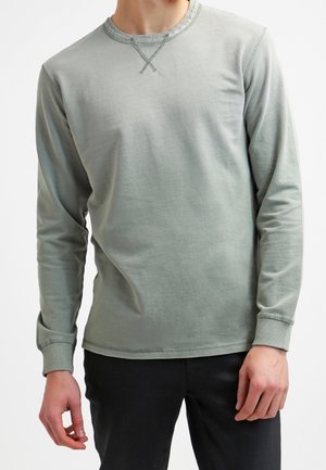 Mand iført en lysegrå langærmet crewneck-trøje med ribstrikkede manchetter og krave, kombineret med sorte bukser.