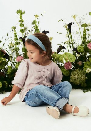 Blusa rosa chiaro con maniche a sbuffo e dettagli a fiocco, abbinata a jeans blu arrotolati e scarpe bianche, seduta di fronte a uno sfondo floreale.