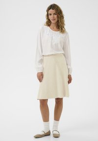 Valge pika varrukaga pluus lillekirjaga, kombineeritud põlvekõrguses beežis seelikuga. Mudel kannab beeże baleriinajalatseid ja valgeid pahkluu sokke.