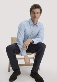 Lichtblauw overhemd met lange mouwen en bruine knopen, gecombineerd met marineblauwe broeken en donkerbruine suède schoenen, zittend op een houten stoel.