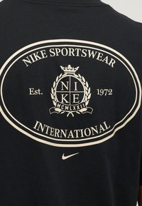 Μαύρο μπλουζάκι πίσω με λευκό λογότυπο Nike Sportswear International, σήμα με το "NIKE" και τα ρωμαϊκά ψηφία "MCMLXXII" και μικρό swoosh από κάτω.