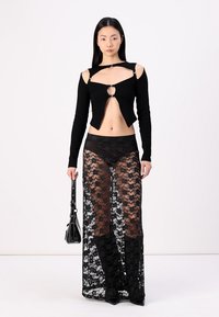 Zwarte crop top met uitsparingen en metalen accenten, gecombineerd met een transparante zwarte kanten maxi rok. Model houdt een kleine zwarte tas vast.