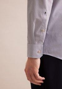 Weißes Hemd mit kleinen blauen und roten runden Mustern. Lange Ärmel mit Knopfmanschetten. Das Material wirkt glatt und leicht.