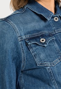 Denimjacka i medelblått med silverdetaljer. Har en spetsig krage, bröstfickor och ljusa sömmar. Strukturerad tyg.