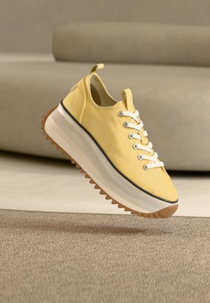 Žlutý plátěný teniska s tlustou bílou podrážkou a hnědým vroubkovaným vzorkem, umístěná nad béžovým kobercem s béžovou pohovkou v pozadí.