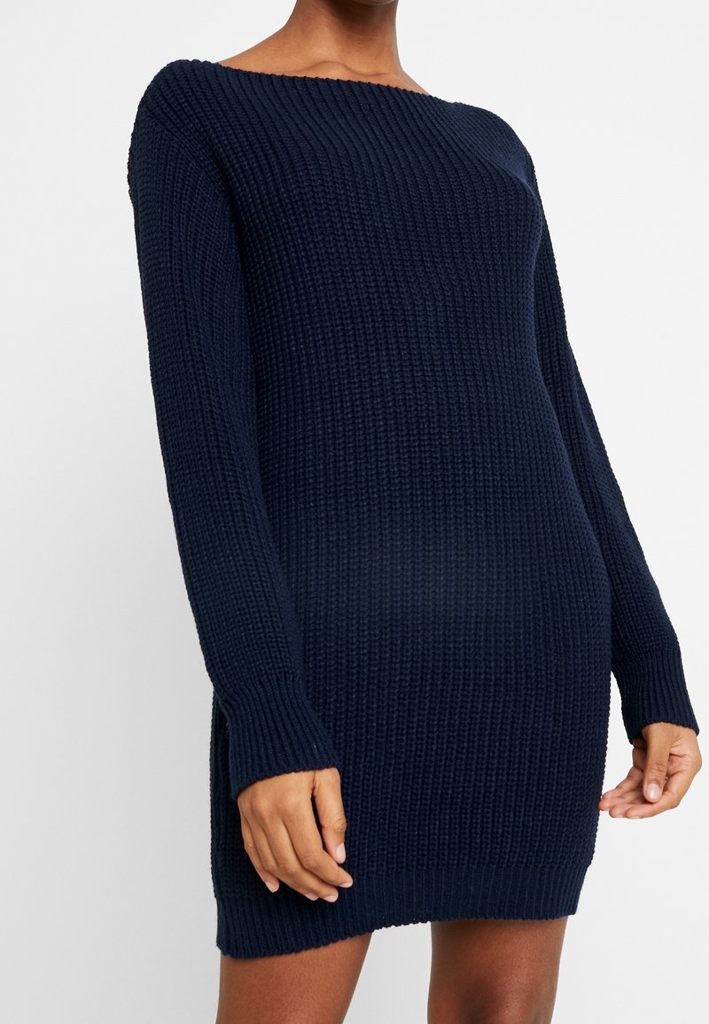 Robe pull en maille bleu marine, présentant une texture côtelée, manches longues et un décolleté épaules dénudées. Longueur mi-cuisse, coupe décontractée.