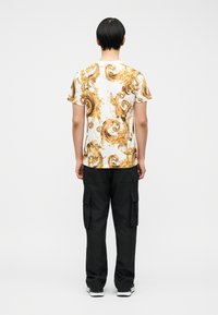  Άντρας με κοντά μαύρα μαλλιά φοράει λευκό μπλουζάκι με χρυσά μπαρόκ σχέδια, μαύρο παντελόνι cargo και αθλητικά παπούτσια, στέκεται με την πλάτη προς τα εμπρός.