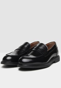 Mocassins en cuir noir avec une finition brillante, dotés d'un détail de bride sur le dessus et d'un bout arrondi. Accents cousus sur la semelle.