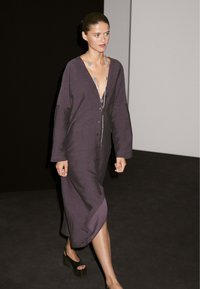 Langes, lila Leinenkleid mit tiefem V-Ausschnitt und Knopfverschluss, das lange Ärmel und seitliche Schlitze aufweist. Die Accessoires umfassen eine silberne Halskette.