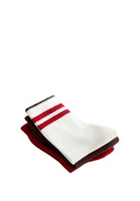 Trois paires de chaussettes pliées empilées, la paire du dessus blanche avec deux bandes rouges et une bordure marron, la paire du milieu marron uni, la paire du bas rouge uni.