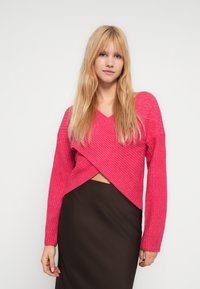 Młoda kobieta o długich blond włosach, ubrana w różowy prążkowany sweter z kopertowym dekoltem oraz wysoką, ciemną spódnicę, stoi na białym tle.