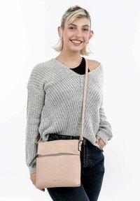 Borsa a tracolla in pelle rosa con chiusura a zip e tasca frontale. La tracolla è regolabile. Indossata in modo informale contro un maglione grigio strutturato.