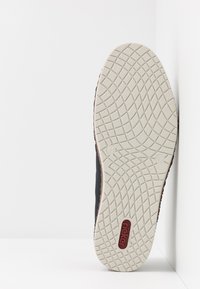 Chaussure noire avec une semelle en caoutchouc texturée présentant un motif de semelle distinctif et une marquage rouge. La semelle est blanche avec un accent marron.