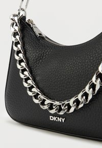 Μαύρη δερμάτινη τσάντα χειρός με υφή, ασημί αλυσίδα και κλείσιμο με φερμουάρ. Το λογότυπο DKNY είναι ανάγλυφο στο μπροστινό μέρος.