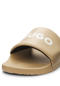 Sandale slide beige à bout ouvert avec semelle texturée et large bride présentant un logo blanc partiel "HUGO".