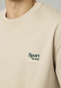 Sweatshirt beige avec un col ras du cou et un texte brodé : "Le sport est ma dépendance" en accents vert et crème. Tissu doux et texturé.