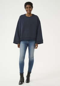 Tumši zilas krāsas pārāk liels džemperis ar teksturētu logo, platām piedurknēm, savienots ar gaiši ziliem šaurajiem džinsiem un melniem potīšu zābakiem. Gluda auduma, ikdienas stils.