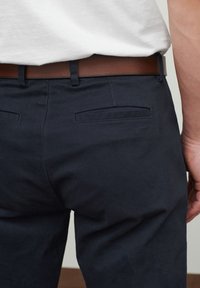 Pantalones azul marino con una textura suave, que cuentan con un cinturón de cuero marrón. La parte trasera tiene dos bolsillos horizontales con bordes cosidos.