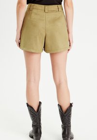 Olive green shorts with a smooth, suede-like texture, featuring a high waist, side pockets, and a folded hem. Black cowboy boots complete the look.