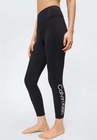 Γυναίκα που φοράει μαύρα κολάν ψηλόμεσα με το λογότυπο Calvin Klein στο κάτω μέρος του ποδιού, στέκεται ξυπόλητη μπροστά σε ένα απλό φόντο.