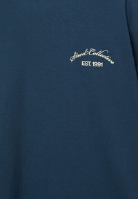 Tissu bleu foncé avec petit texte cursif de couleur crème "Stud Collective" et "EST. 1991" imprimé en haut à gauche.